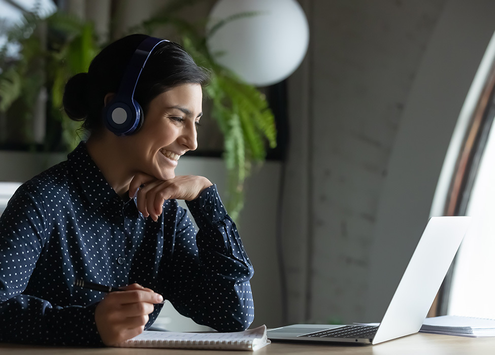 Business Technology: Person with headphones on smiles as they work on their laptop while also taking notes in a notebook