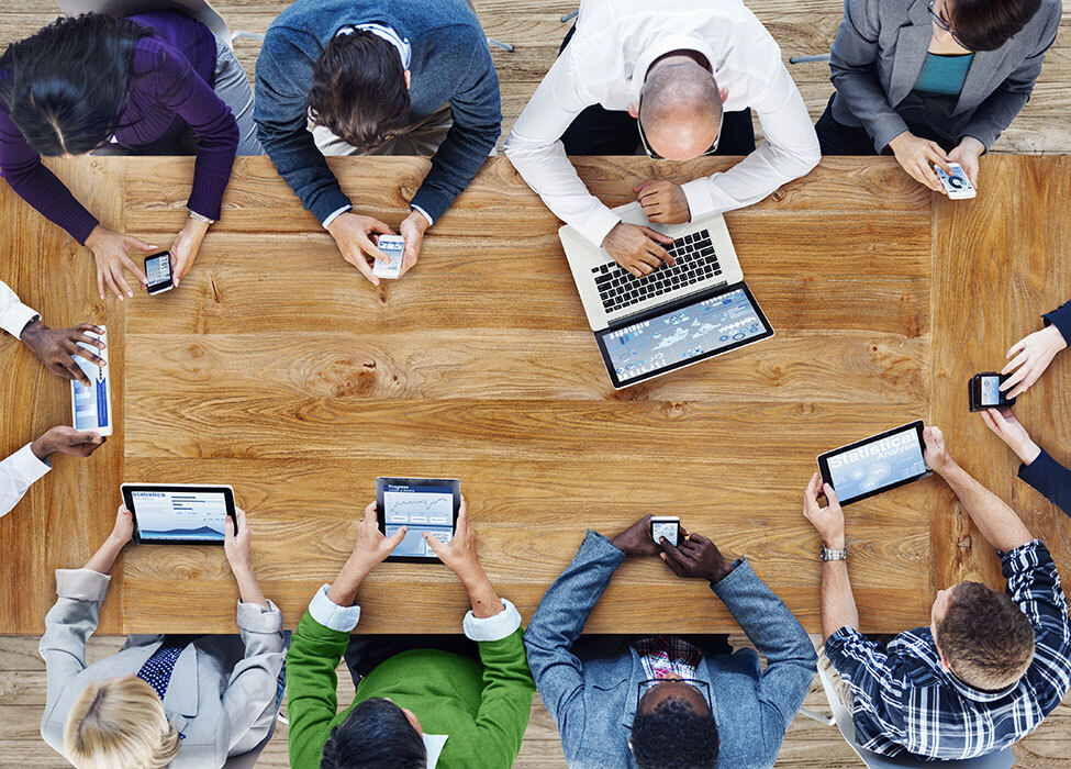 Smart Office Technology | View from above of multiple people sitting at a table working on laptops, tablets, and phones