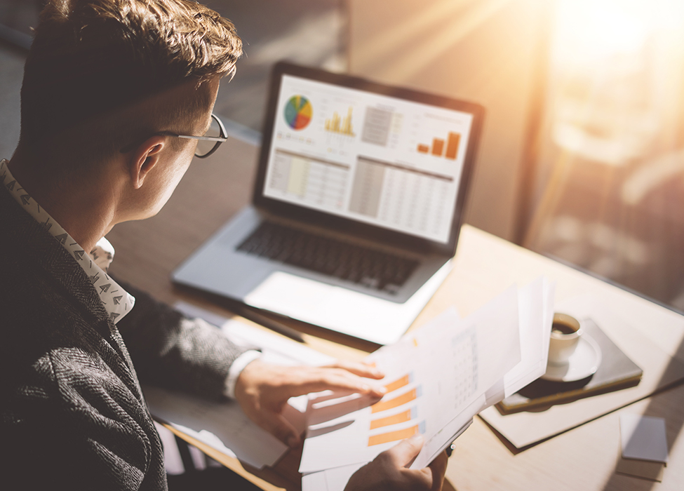 KYC Automation | Person holding papers with graphs on them and comparing them to graphs on their laptop screen