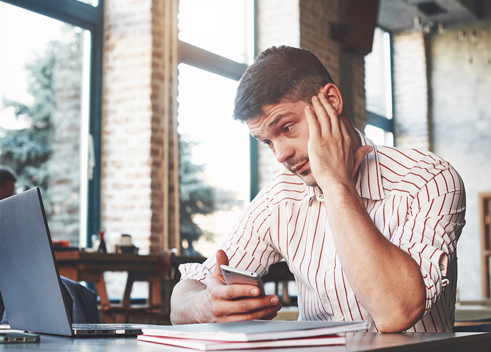 Man rubs his head in frustration while holding his phone and looking at his laptop