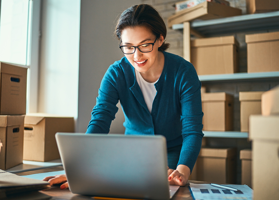 Smart Technology | woman in office with a lot of boxes smiling and working on the computer 