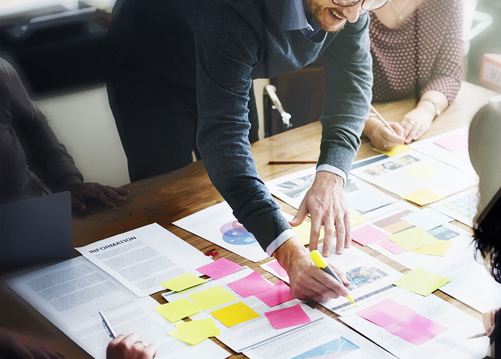 Integrated Marketing Strategy | people meeting and looking at multiple papers scattered on table. One person highlights information on one of the papers.